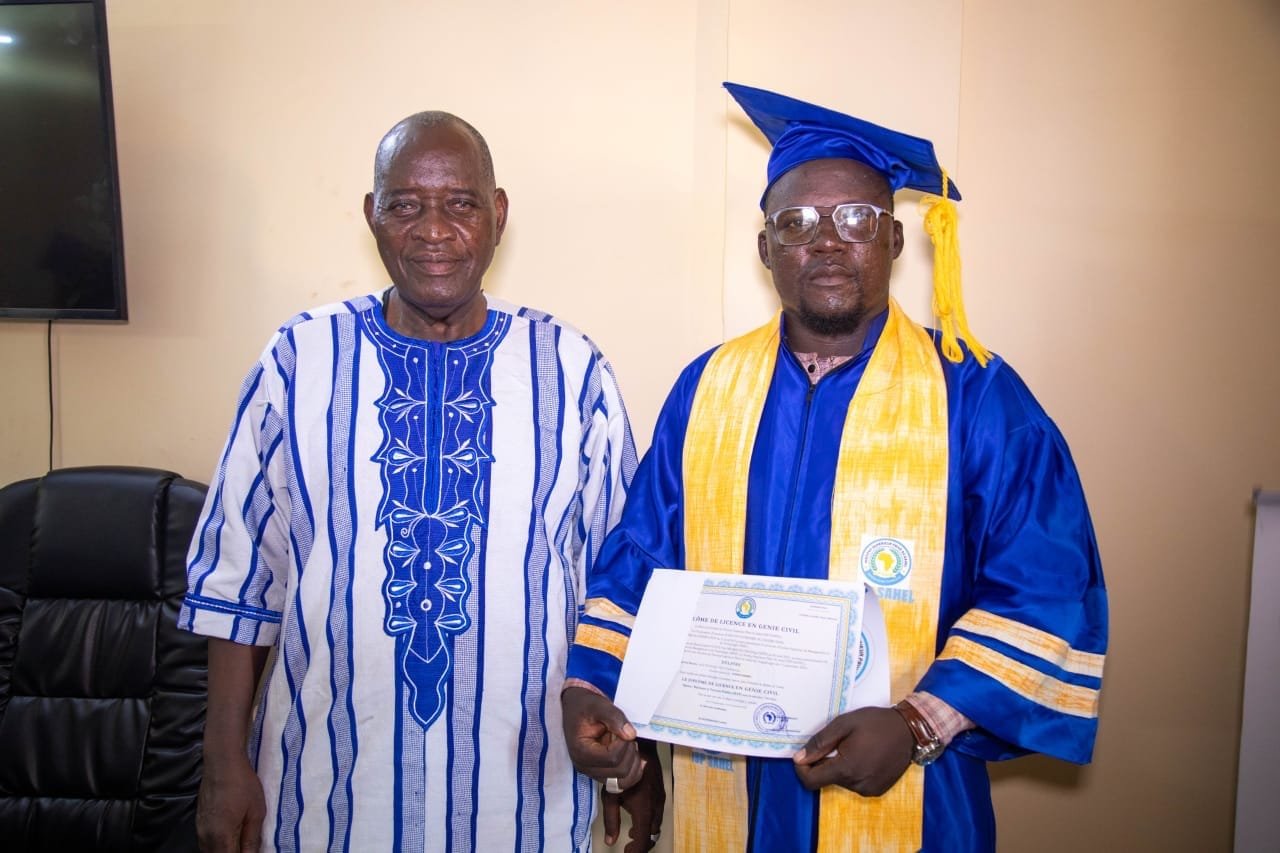 Un diplômé en toge bleue présente son diplôme de Licence en Génie civil aux côtés d'un aîné en boubou
