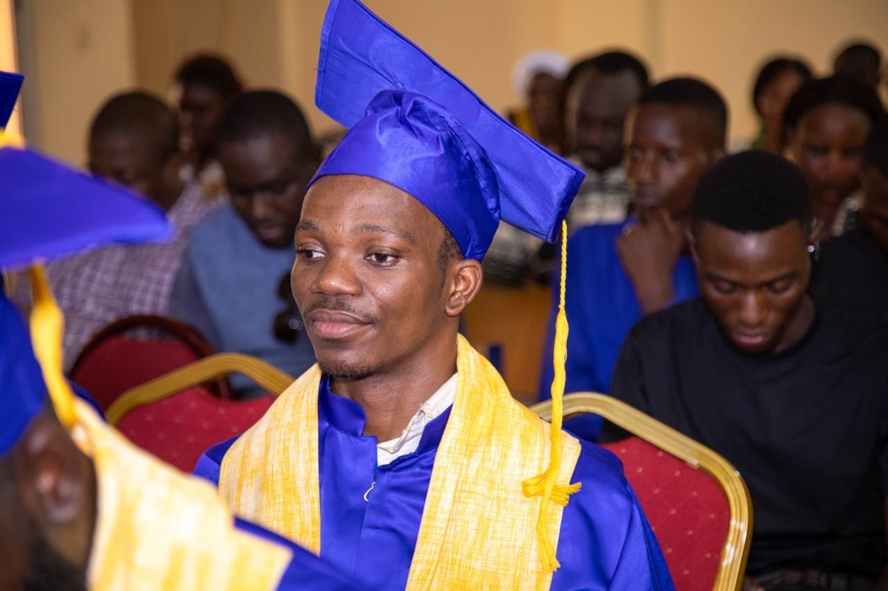 Un diplômé en toge bleue et étole jaune, souriant, assis dans la salle