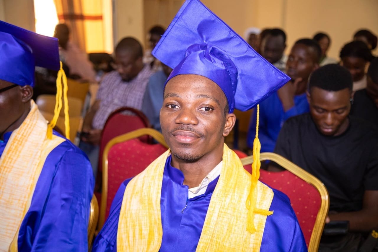 Un diplômé souriant en toge bleue et étole jaune, assis dans la salle