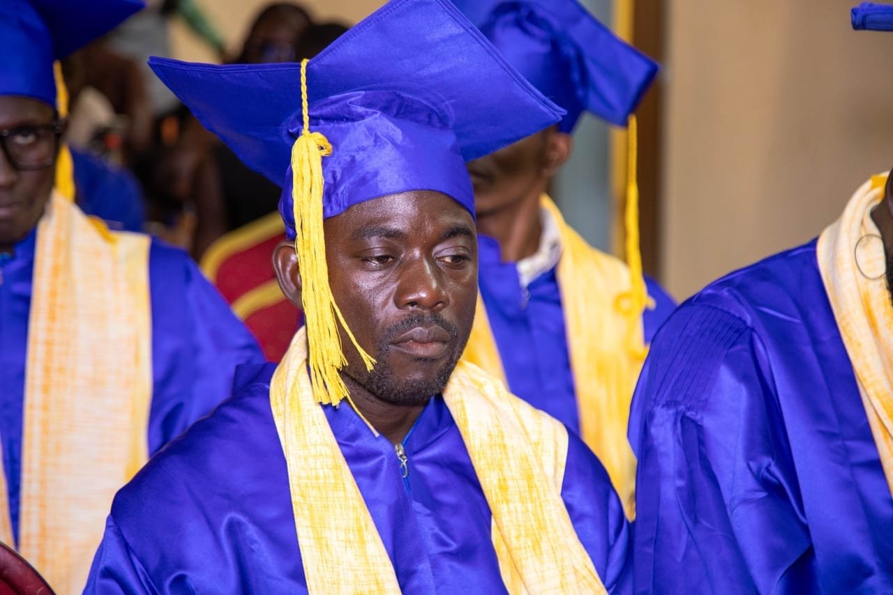 Un diplômé en toge bleue et étole jaune, le regard baissé, entouré d'autres diplômés